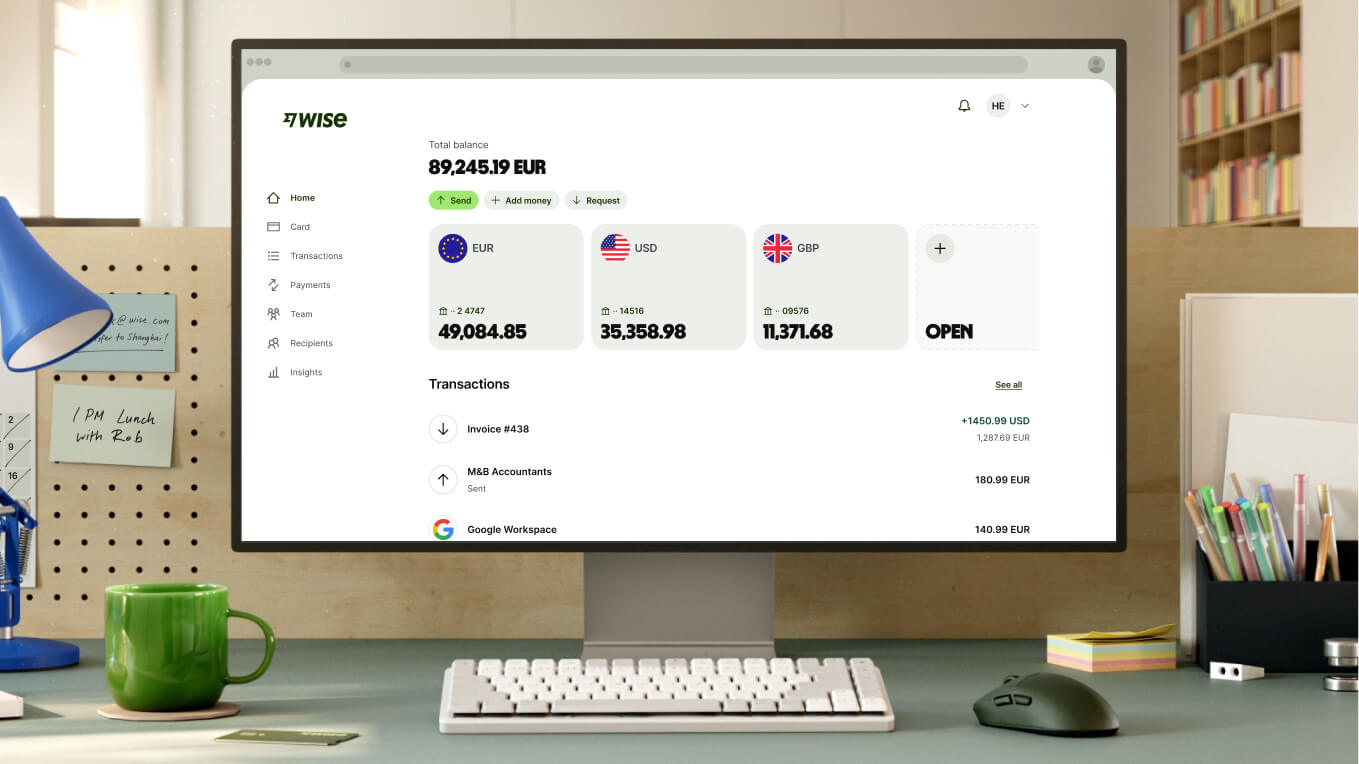 A close-up shot of a modern workspace shows a white computer monitor displaying the Wise dashboard. The screen shows a total balance of "89,245.19 EUR" and three main currency accounts: EUR with "49,084.85," USD with "35,358.98," and GBP with "11,371.68," followed by an option to open a new account. Recent transactions are listed below. In front of the monitor is a compact white keyboard, a dark grey mouse, and a bright green ceramic mug.