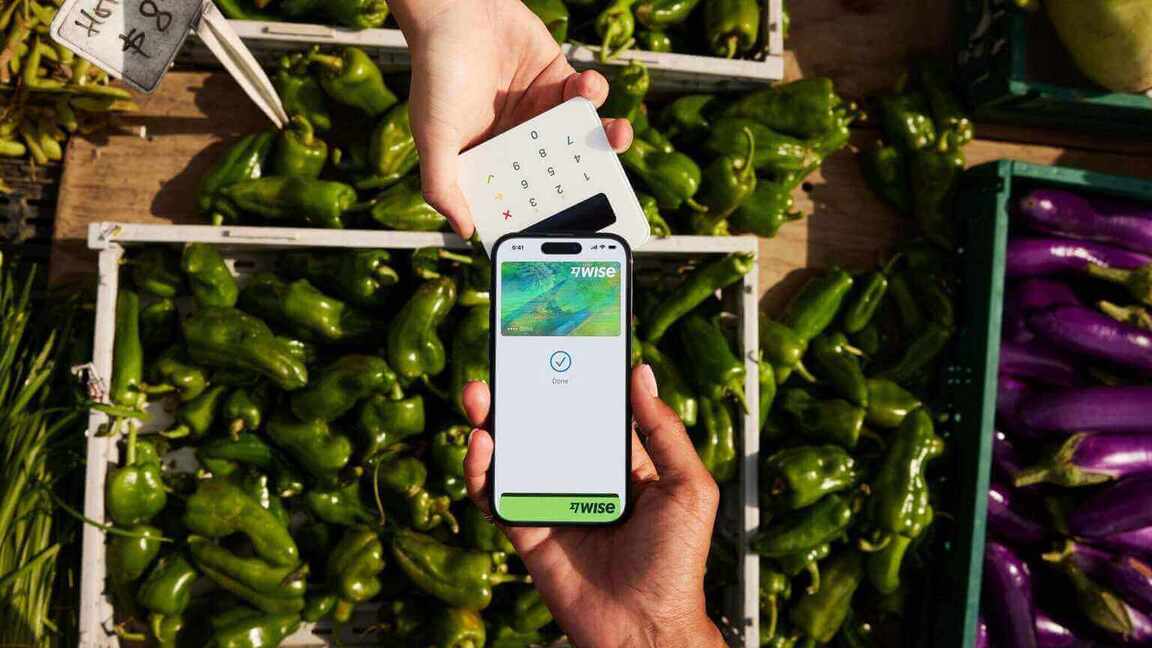 Two hands over a box of peppers, one holding a phone showing the Wise digital card, the other holding a payment tool