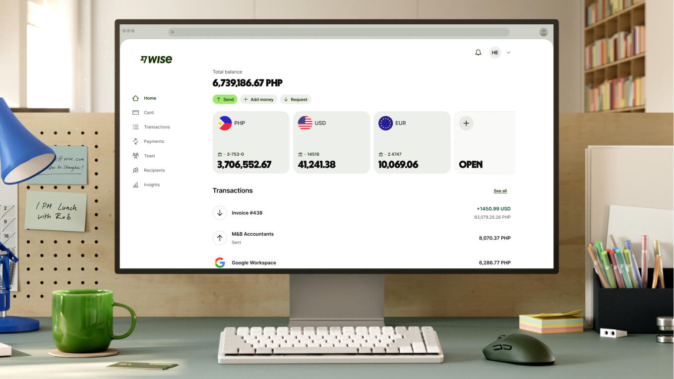 A close-up shot of a modern workspace shows a white computer monitor displaying the Wise dashboard. The screen shows a total balance of "6,739,186.67 PHP" and three main currency accounts: PHP with "3,706,552.67," USD with "41,241.38," and EUR with "10,069.06," followed by an option to open a new account. Recent transactions are listed below. In front of the monitor is a compact white keyboard, a dark grey mouse, and a bright green ceramic mug.